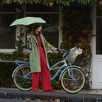 A person standing outdoors next to a light blue bicycle, wearing a long sage green coat over a red dress and red tie, holding a Blunt Umbrella Neo Sage Green.